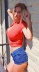 Woman leaning against a light brick wall in a red cropped top and blue denim shorts, posing with one hand in her hair and looking at the camera nearby.